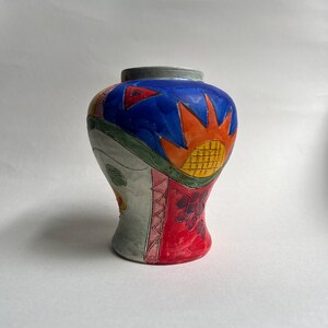 Może przedstawiać: Ceramiczny wazon z żywym, ręcznie malowanym wzorem, posiadający niebieską górną część, czerwoną dolną część oraz szary panel boczny. Powierzchnia ozdobiona jest kolorowymi motywami graficznymi, w tym żółtym słońcem i czerwonymi trójkątami.