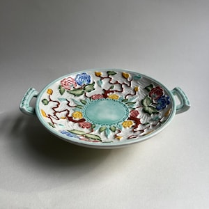 Peut inclure: Plat de service décoratif en céramique avec un motif floral. Le plat présente un bord et des poignées bleu clair, avec un intérieur blanc orné de fleurs colorées rouges, bleues et jaunes. Le plat mesure environ 30 cm de diamètre.