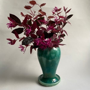 May include: A green ceramic vase with a flared base holds a bouquet of deep purple flowers with pink blooms.