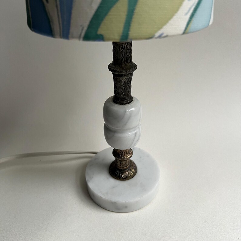 May include: A small table lamp with a white marble base and a patterned lampshade. The lamp features a decorative metal stem and a cylindrical marble body. The lampshade has a blue, green, and white design.