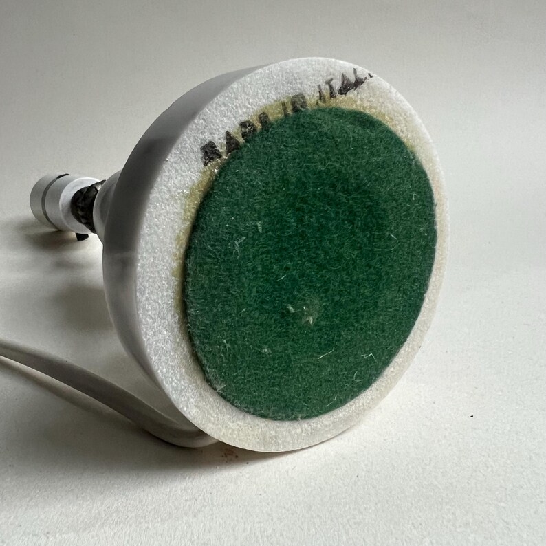 May include: A small, white lamp base with a green felt bottom. The base has a circular shape with a white cord and the text "MADE IN ITALY". The lamp's design is simple and modern.