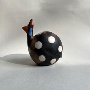 Puede incluir: Figura de pájaro de cerámica con cuerpo redondeado y cuello largo. El pájaro es principalmente negro con grandes lunares blancos. La cabeza y el cuello presentan detalles marrones, rojos y azules. El objeto es decorativo.