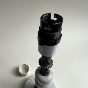 May include: A lamp base with a black and white bulb socket, a decorative metal stem, and a marble base. A small white cap sits to the side. The lamp base is designed for a light fixture.