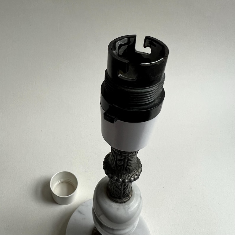 May include: A lamp base with a black and white bulb socket, a decorative metal stem, and a marble base. A small white cap sits to the side. The lamp base is designed for a light fixture.