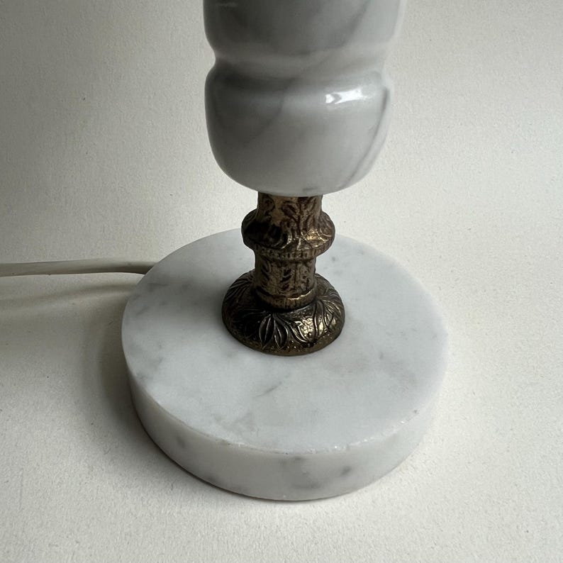 May include: A small, decorative lamp with a white marble base and a white marble lamp shade. The lamp has a bronze-colored, ornate base and a power cord. The lamp is a decorative home accent.