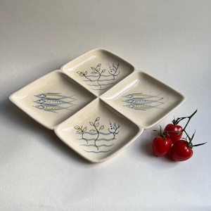 May include: A set of four off-white ceramic serving dishes arranged in a diamond shape. Each dish features a unique design of stylized fish or aquatic plants in blue, yellow, and black. Red tomatoes are on the right.