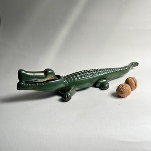 May include: A dark green alligator-shaped nutcracker with a textured, scaly back and a hinged jaw mechanism. The alligator is positioned horizontally with its mouth slightly open, accompanied by two brown, textured walnuts placed nearby.
