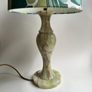 May include: A table lamp with a polished green stone base featuring natural veining and a curved, hourglass-shaped stem. The lamp is topped with a drum-style lampshade decorated with a marbled pattern in shades of green, blue, and white.
