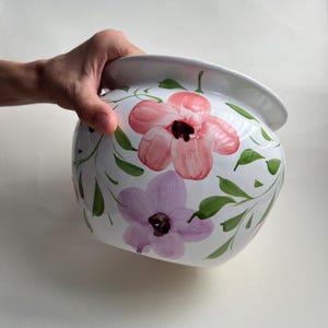 May include: A white ceramic bowl featuring a hand-painted floral design. The exterior displays large pink and light purple flowers with dark centres, accented by green leaves and thin vines. The bowl has a smooth, glossy finish and a wide, rounded shape.
