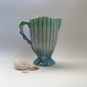 Könnte beinhalten: Eine Keramikkrug mit Muscheldesign in Blau- und Grüntönen. Der Griff und der Sockel weisen ein wellenartiges Design auf. Zwei Muscheln und ein Sanddollar befinden sich neben dem Krug.