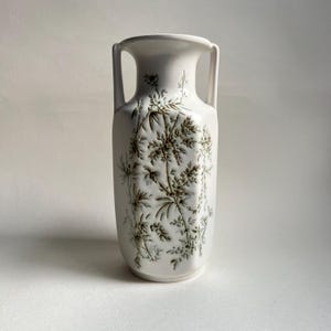 May include: White ceramic vase with two handles and a botanical design in shades of brown and green. The vase has a rectangular shape with a flared top and is suitable for displaying flowers or as a decorative piece.