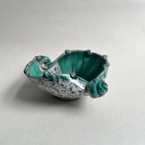 May include: A ceramic bowl featuring a deep teal interior glaze that transitions into a white and grey speckled exterior. The rim is uneven and wavy, accented with small, rounded teal protrusions that extend outward from the bowl's edge.