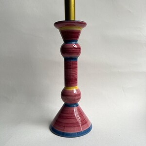 May include: A decorative candlestick holder with a striking design. The ceramic piece has a burgundy body with horizontal brushstrokes, accented by yellow and blue bands. The top has a brass fitting.