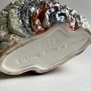 May include: Close-up of the bottom of a ceramic figurine. The figurine is brown, green, and white with a textured surface. The bottom of the figurine is white with the words "STAVAO MADE IN ENGLAND" and the number "3530" printed on it.