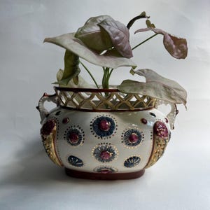 May include: A white ceramic planter with two handles and a lattice rim, decorated with repeating dark blue and magenta floral motifs outlined in gold. A plant with light pink and green variegated leaves grows from the centre of the vessel.