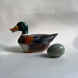 May include: A ceramic duck-shaped trinket box with a green head, orange and brown body, and a yellow beak. Beside it is a small, oval, speckled green stone. The duck box is a decorative item.