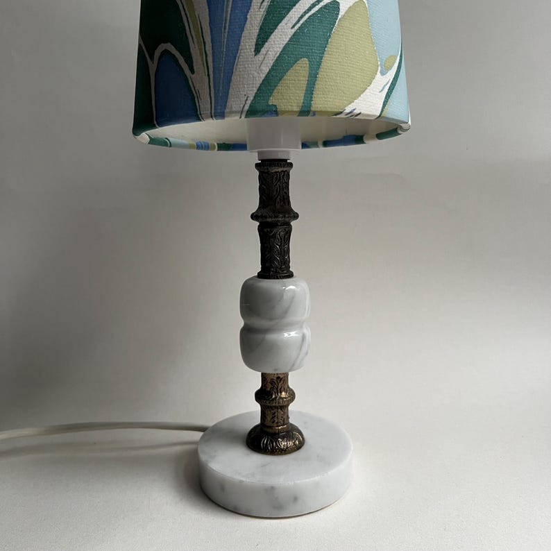 May include: A small table lamp with a marble base and a patterned lampshade. The lamp features a white and gray marble body with bronze-colored metal accents. The lampshade has a blue, green, and white abstract design.