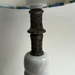 May include: A decorative table lamp with a white marble base and a dark, ornate metal column. The lamp shade is visible at the top, featuring a pattern of blue, green, and white stripes. The lamp is designed for indoor use.