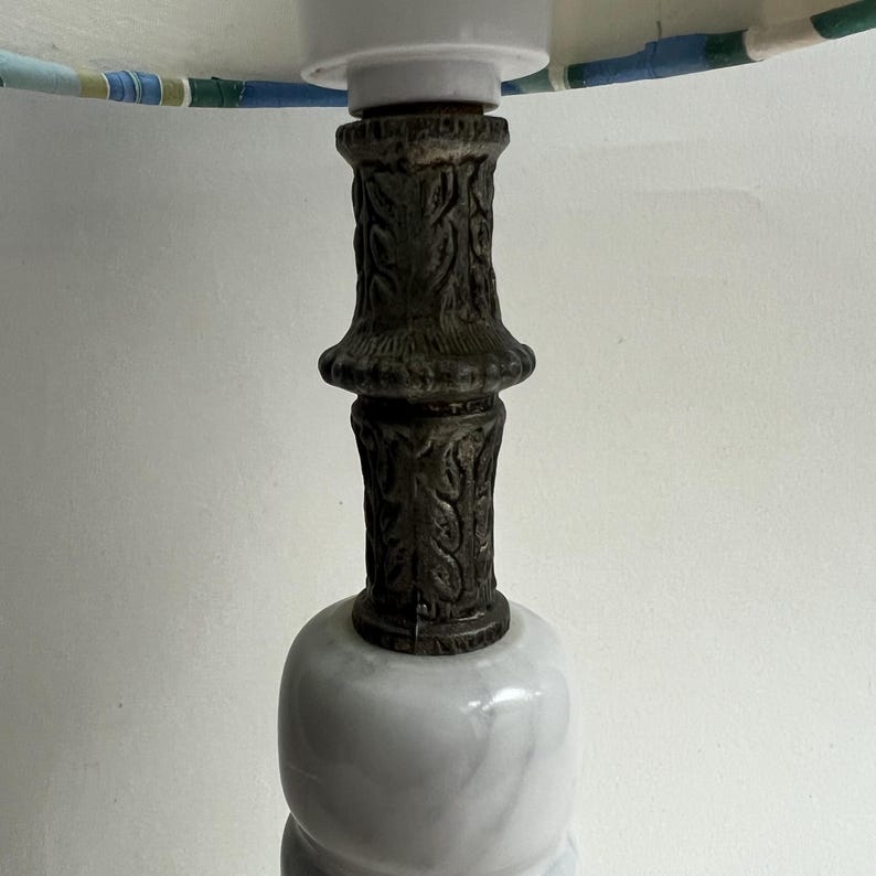 May include: A decorative table lamp with a white marble base and a dark, ornate metal column. The lamp shade is visible at the top, featuring a pattern of blue, green, and white stripes. The lamp is designed for indoor use.