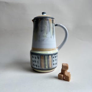 May include: A ceramic coffee pot with a lid, handle, and spout. The pot has a white body with a decorative band of black, yellow, and brown vertical stripes. A small stack of brown sugar cubes sits beside the pot.