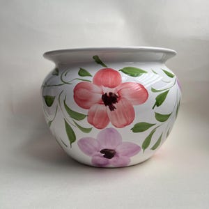 Peut inclure: Un cache-pot en céramique blanche avec un corps arrondi et un large rebord. L'extérieur présente des motifs floraux peints à la main dans des tons de rose et de violet, agrémentés de feuilles et de vignes vertes sur la surface blanche brillante.