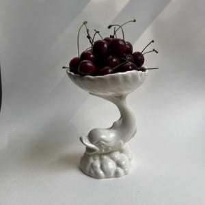 May include: A white ceramic pedestal bowl featuring a sculpted dolphin base rising from a cloud-like foundation. The bowl is filled with a cluster of dark red cherries with thin green stems, creating a contrast between the fruit and the white dish.