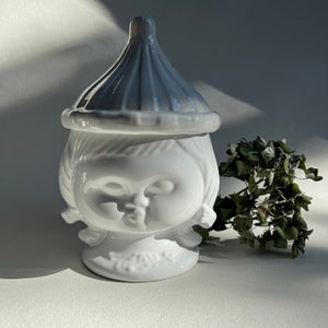 May include: A white ceramic jar with a lid, depicting a stylized face. The lid is light grey, and the jar has a whimsical, doll-like appearance. A sprig of dried greenery is next to the jar. The jar is likely for decorative purposes.