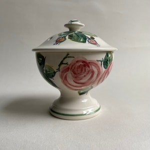 May include: A white ceramic lidded bowl with a pedestal base. The bowl and lid are decorated with pink roses, green leaves, and buds. The rim of the base has a green stripe.