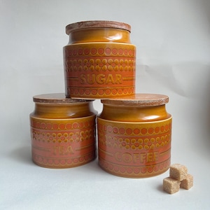 May include: Three ceramic kitchen canisters in shades of orange and brown, labeled "Sugar", "Tea", and "Coffee". Each canister has a wooden lid and a decorative floral pattern. Three sugar cubes are in the foreground.