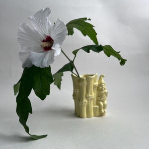 Puede incluir: Un florero de cerámica amarillo pálido con forma de tallos de bambú y una pequeña figura de gnomo adherida al costado. Una sola flor de hibisco blanca grande con un centro rojo oscuro y hojas verdes está colocada dentro del florero.