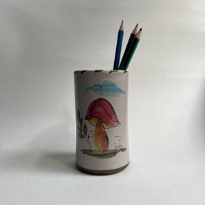 Op de afbeelding: Een cilindervormige keramische potloodhouder met een lichtbeige basiskleur. De buitenkant is voorzien van een handgeschilderde illustratie van een paddenstoel met een roze-rode hoed en oranje steel, een kleine blauwe wolk en groen blad. Drie potloden staan erin.