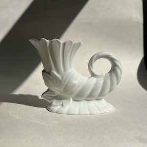 Puede incluir: Un jarrón de cerámica blanco en forma de cornucopia, con un borde acanalado y un asa en forma de bucle. El jarrón tiene un diseño detallado y texturizado con patrones de hojas y espirales. La base tiene forma redondeada y de pedestal. El jarrón mide unos 15 cm de alto.