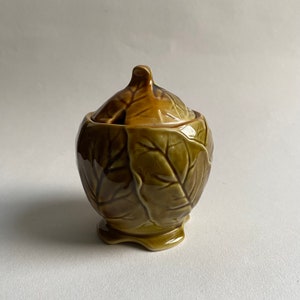 Può includere: Una zuccheriera in ceramica con coperchio, a forma di cavolo con foglie verdi e marroni.