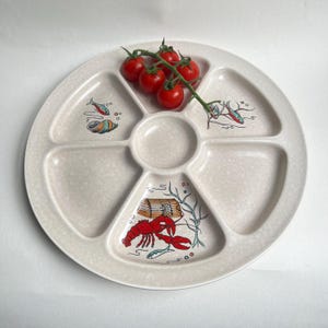 May include: A beige, speckled, divided plate with a central well and five compartments. Each section is decorated with marine illustrations, including a red lobster, fish, and a seashell. A cluster of red cherry tomatoes rests on the plate.