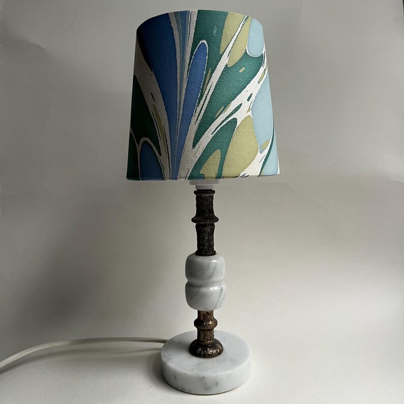 May include: A decorative table lamp with a marble base and a patterned lampshade. The lampshade features a swirling design in shades of blue, green, and yellow. The lamp's stem has a dark, ornate design. The lamp is a stylish home decor item.
