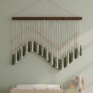 May include: A macrame wall hanging with a dark brown wooden rod and white cords. The design features a zig-zag pattern of wooden beads and green and cream-colored tassels. The wall is painted a light green color.