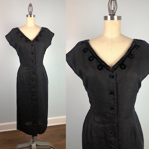 May include: A vintage black dress with a V-neckline and cap sleeves. The dress features a row of buttons down the front and decorative trim around the neckline. The dress is displayed on a mannequin.