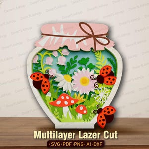 Può includere: Un design multistrato tagliato al laser con un barattolo pieno di fiori, coccinelle e funghi. Il barattolo ha un coperchio rosa legato con un fiocco marrone. Il testo "Multilayer Lazer Cut" è in basso.
