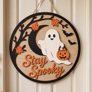 May include: A circular wooden wall sign featuring a white ghost holding an orange jack-o'-lantern bucket. The design includes a black crescent moon, a black tree branch, orange maple leaves, two black bats, and the orange text Stay Spooky.