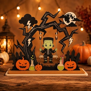 Pu&ograve; includere: Una decorazione di Halloween in legno a strati raffigurante un personaggio verde in stile Frankenstein seduto su un'altalena sotto un albero nero con fulmini e luna piena. La scena include due zucche arancioni e due becher in vetro.