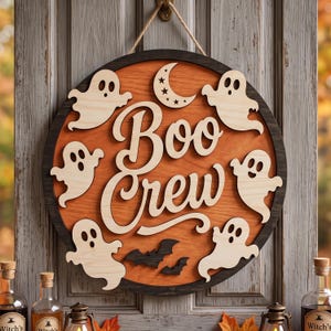 May include: A circular wooden wall sign featuring a dark frame and an orange centre. The text "Boo Crew" is written in large, light-coloured script. The sign is decorated with five light-coloured ghost cutouts, two black bats, and a crescent moon.