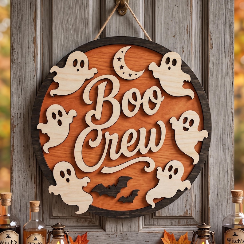 May include: A circular wooden wall sign featuring a dark frame and an orange centre. The text "Boo Crew" is written in large, light-coloured script. The sign is decorated with five light-coloured ghost cutouts, two black bats, and a crescent moon.