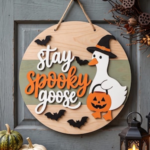 May include: A round wooden wall sign featuring a white goose wearing a black witch hat and holding an orange jack-o'-lantern bucket. The sign displays the text "stay spooky goose" in white and orange lettering, accented by small black bats.