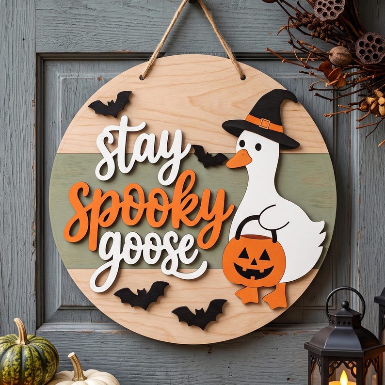 May include: A round wooden wall sign featuring a white goose wearing a black witch hat and holding an orange jack-o'-lantern bucket. The sign displays the text "stay spooky goose" in white and orange lettering, accented by small black bats.