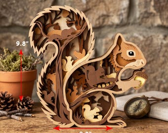 Op de afbeelding: Een meerlaags houten eekhoornsculptuur in diverse bruintinten, 24,9 cm hoog en 23,6 cm breed. Het ontwerp bevat gedetailleerde uitsparingen van eekhoorns en bomen, waarbij de centrale eekhoorn een eikel vasthoudt in de voorpoten.