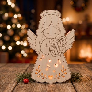 May include: A light-colored wooden cutout of an angel with a halo, holding a harp. The angel's dress features cut-out shapes of musical notes, stars, and a heart, which are illuminated from within, casting a warm glow through the wooden surface.