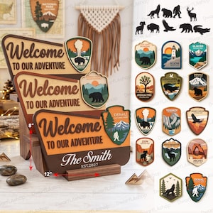 Puede incluir: Tres letreros de madera en capas con el texto Welcome to our adventure, The Smith y EST.2027. Una herramienta de medición roja indica un ancho de 30,48 cm y una altura de 20,32 cm. Los letreros presentan emblemas de parques nacionales.