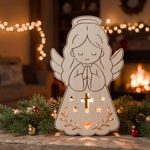 May include: A light-colored wooden angel figurine with a halo, featuring cut-out details including a central cross, four stars, and a heart shape at the base. The angel has closed eyes and hands pressed together in a prayer position, with light glowing from within.