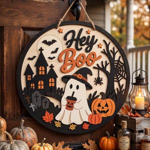 May include: A circular wooden door sign featuring the text Hey Boo in black and orange. The design includes a white ghost wearing a witch hat and holding a cup, a black haunted house, orange pumpkins, bats, a spider, and autumn leaves.