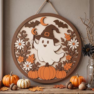 May include: A circular wooden wall sign featuring a layered design of a white ghost wearing a brown witch hat, holding a crescent moon wand. The scene includes orange pumpkins, brown bats, and white flowers with orange centres, all framed by a dark brown border.